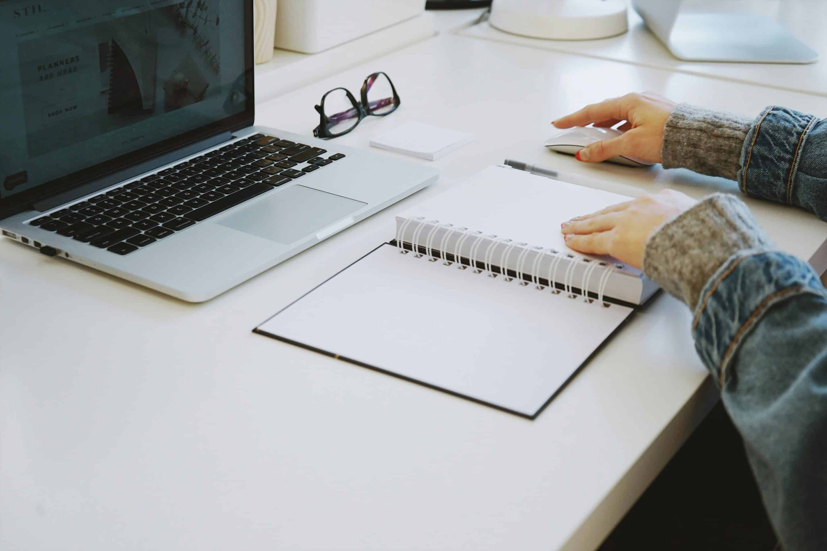 Hands on notebook at a desk with laptop and glasses.